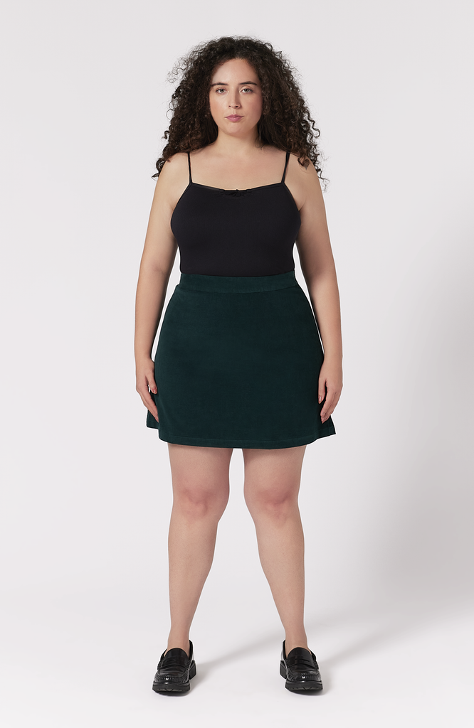 The Cord Mini Skirt by Aam The Label is designed to sit snug at the high waist with a roomy fit for full hips and thighs. Featuring a small, center back elastic for comfort fit at the waist, and hidden zipper closure for a seamless finish. Made from a soft 97% cotton corduroy with 3% stretch. Shown here is the full frontal view of the skirt in a deep, forest green.