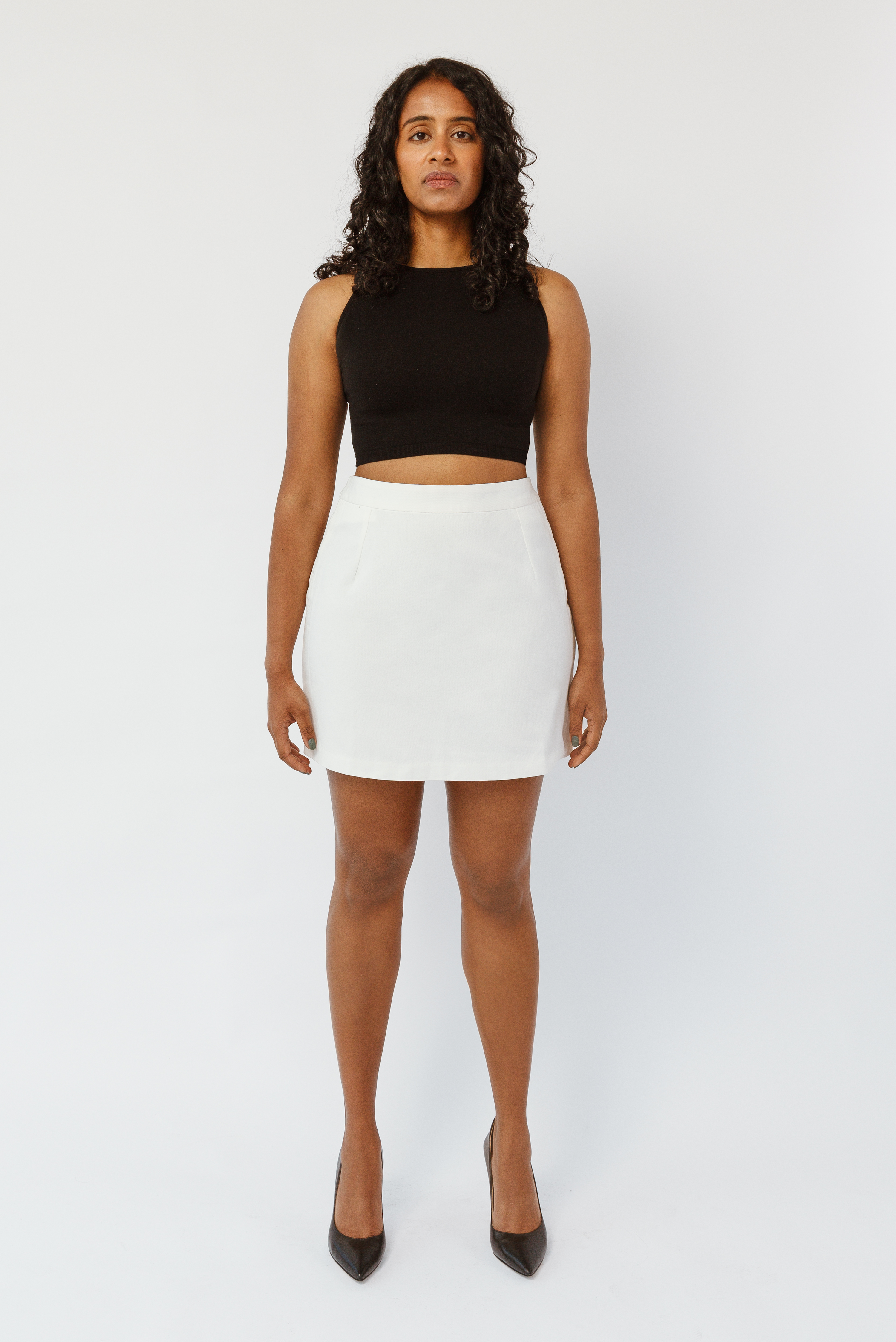 The Mini Skirt by Aam is designed for women with full hips and thighs. It's sit snug at the high waist with roomy fit through the seat for curvy bottom shapes. This image shows the front view of The Mini Skirt in white. It is shown on a model who is 5’7” and wearing a size Small. The skirt has been transparency tested across skin tones and is opaque.